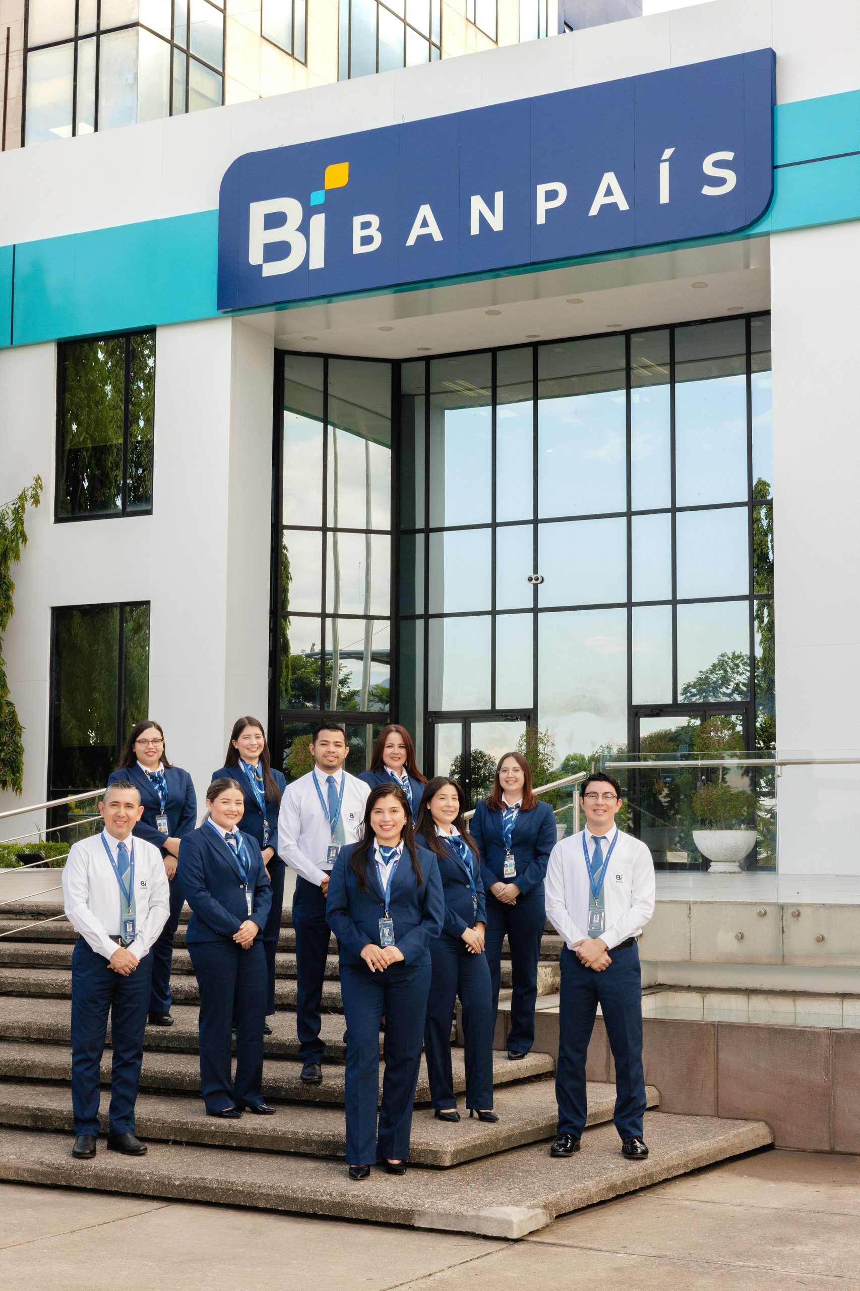 Equipo de Banpaís con el uniforme nuevo en las gradas de la agencia, bajo el rótulo Bi BANPAÍS